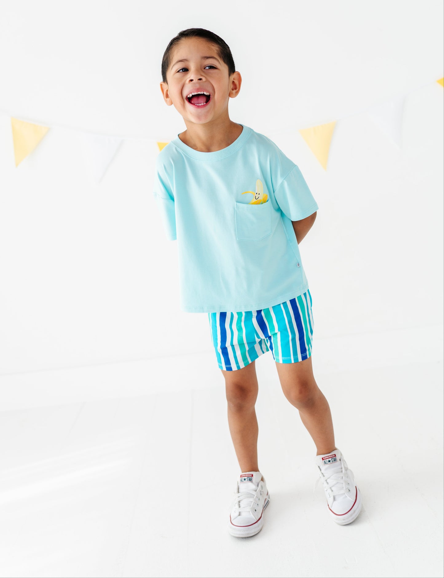 A young boy smiling and posing in a light blue t-shirt with a small yellow pocket graphic and matching blue striped shorts, paired with white sneakers, against a white studio background with bunting.