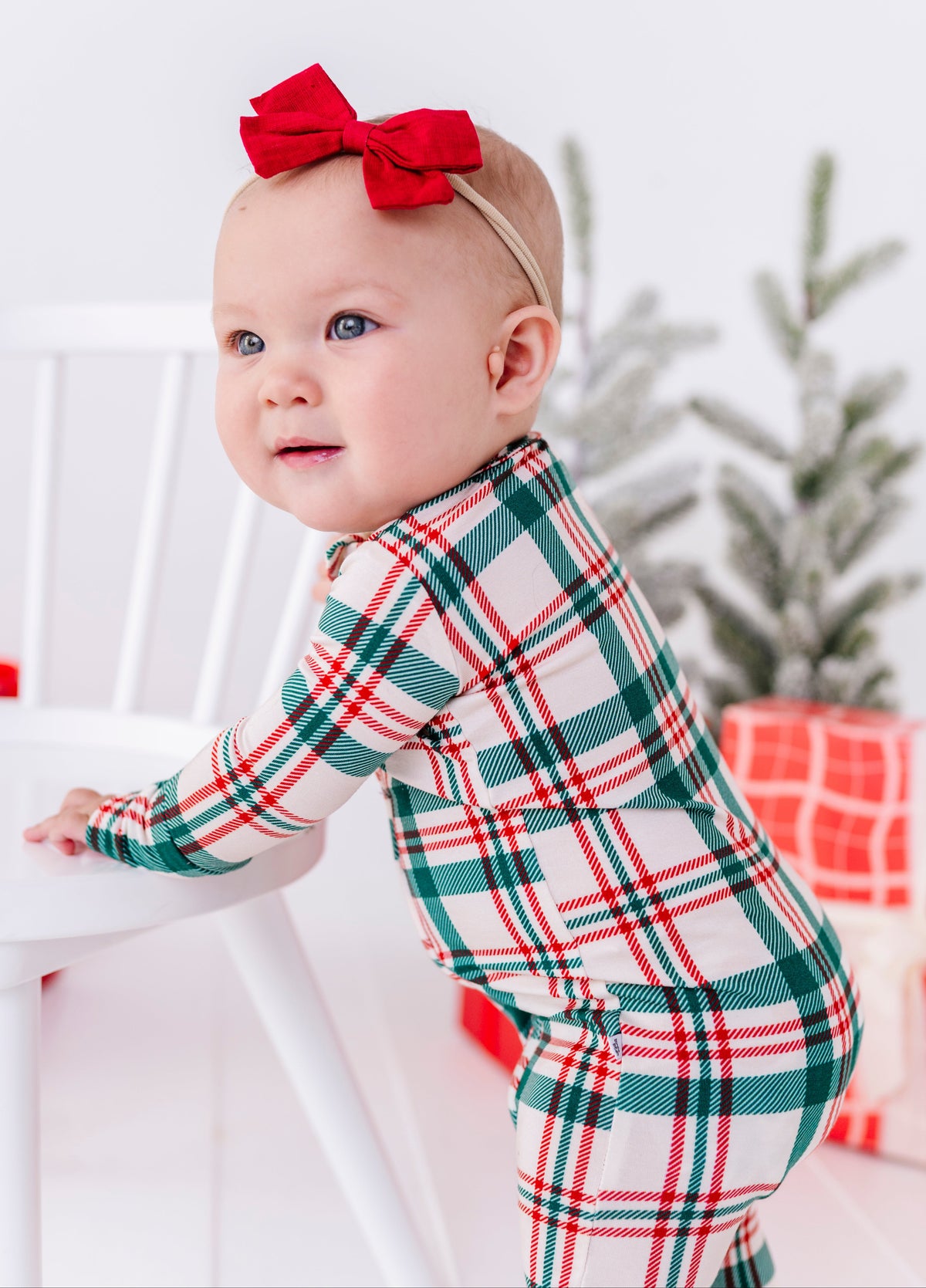 A baby wearing a festive green, red, and white plaid long-sleeve romper and a matching red bow headband, leaning on a white chair in a holiday-themed setting.