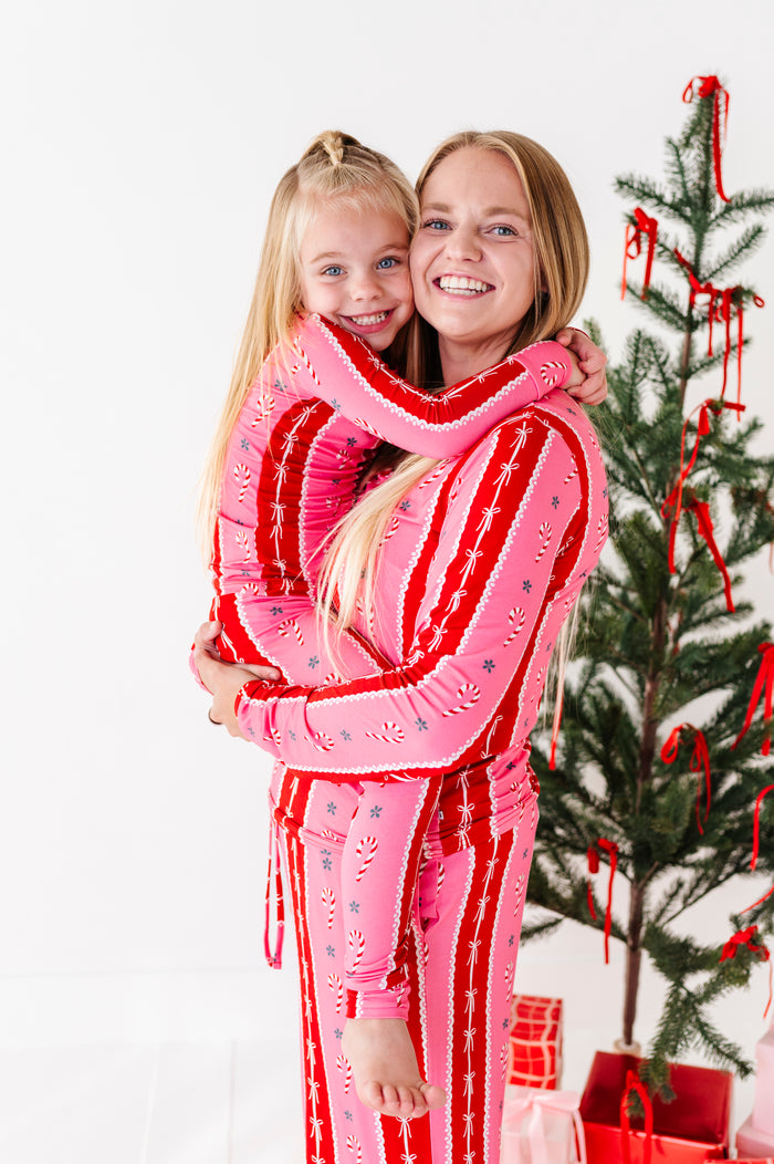 A woman and a young girl hugging while wearing matching pink and red holiday-themed bamboo pajama sets, posed in front of a decorated Christmas tree on a white studio background.