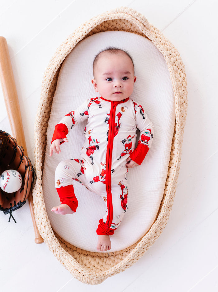 A baby boy wearing a bamboo convertible romper featuring various Santa Clauses throwing baseballs and playing baseballs. The baby is posed with a leather glove, baseball, and wooden baseball bat