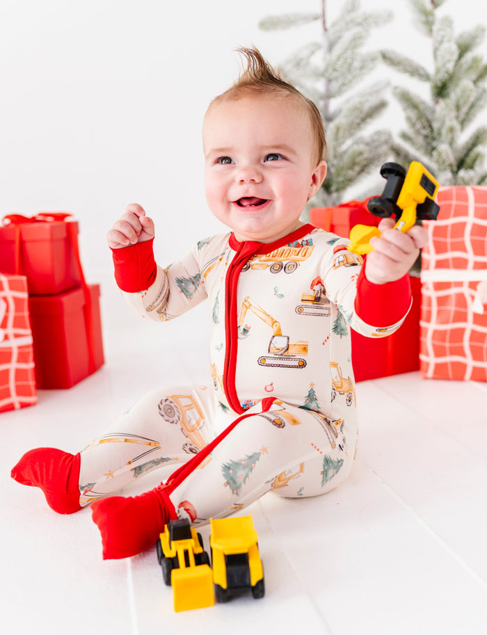 A baby boy wearing a construction-themed bamboo zip romper and playing with construction trucks. He's sitting in front of a Christmas tree and a pile of red wrapped presents