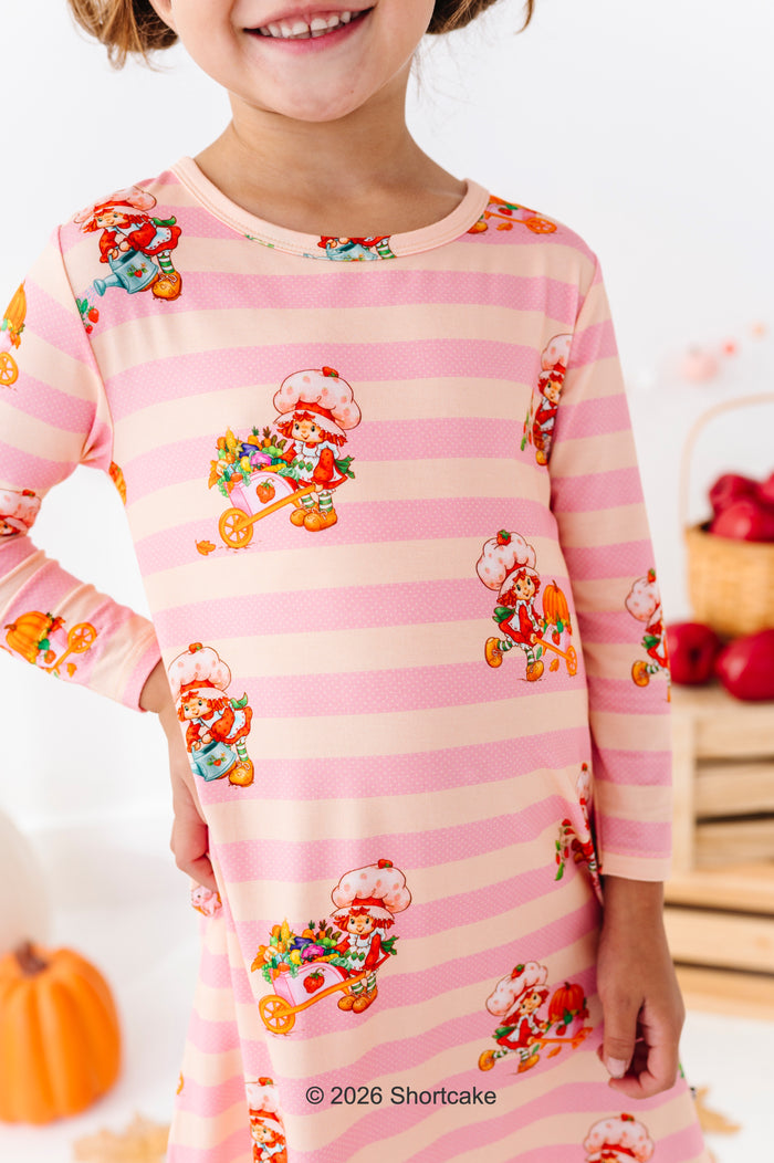 A child wearing a pink and cream striped long-sleeve pajama top with a cartoon character pattern, shown in a cozy indoor setting with a pumpkin and basket in the background.