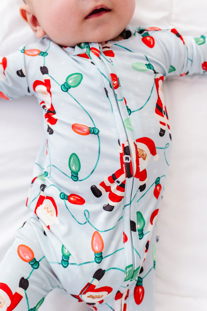 A close-up detail shot of a little boy wearing a blue bamboo convertible romper featuring Santa Claus hanging from multi-colored Christmas lights