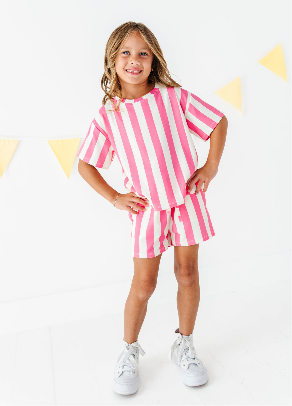A young girl stands smiling in a pink and white vertically striped short-sleeve t-shirt and matching striped shorts, paired with white sneakers, against a white background with yellow bunting.