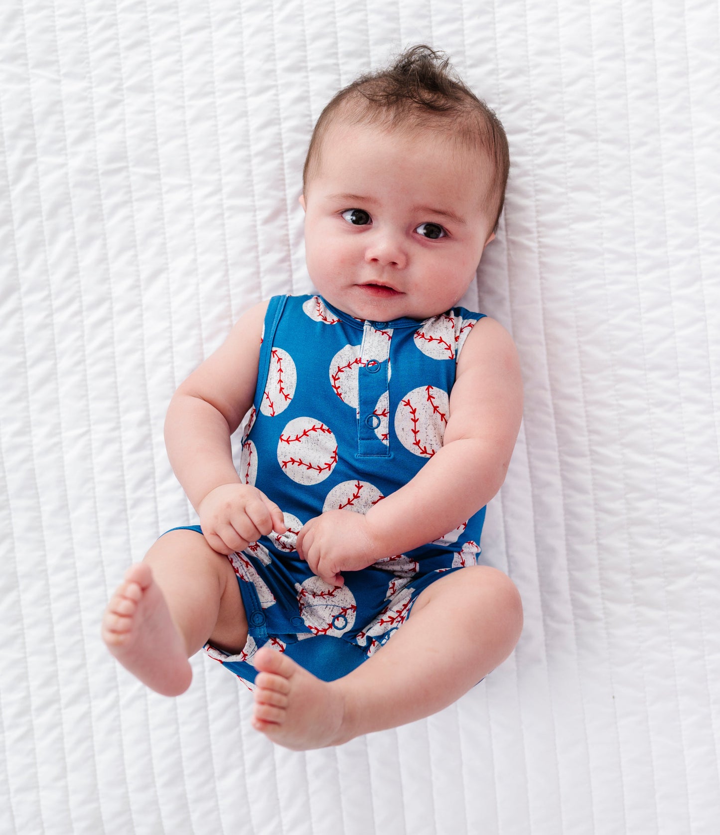 Baby wearing a blue romper with baseball pattern on a white textured blanket