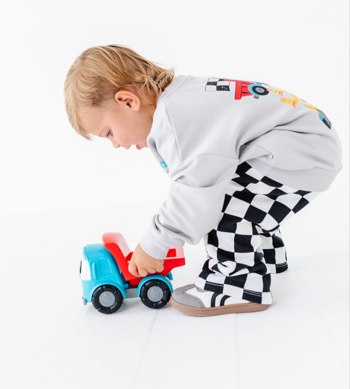 A toddler wearing a light gray sweatshirt and black-and-white checkered pants pushes a blue and red toy dump truck on a clean white floor against a white background.