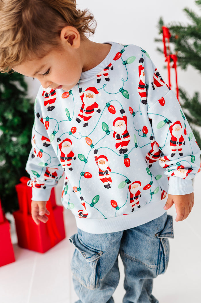 A young child wearing a white long-sleeve crewneck sweatshirt with a festive Christmas print of Santa Claus and holiday lights, paired with blue jeans, standing in front of a decorated Christmas tree and wrapped gifts.