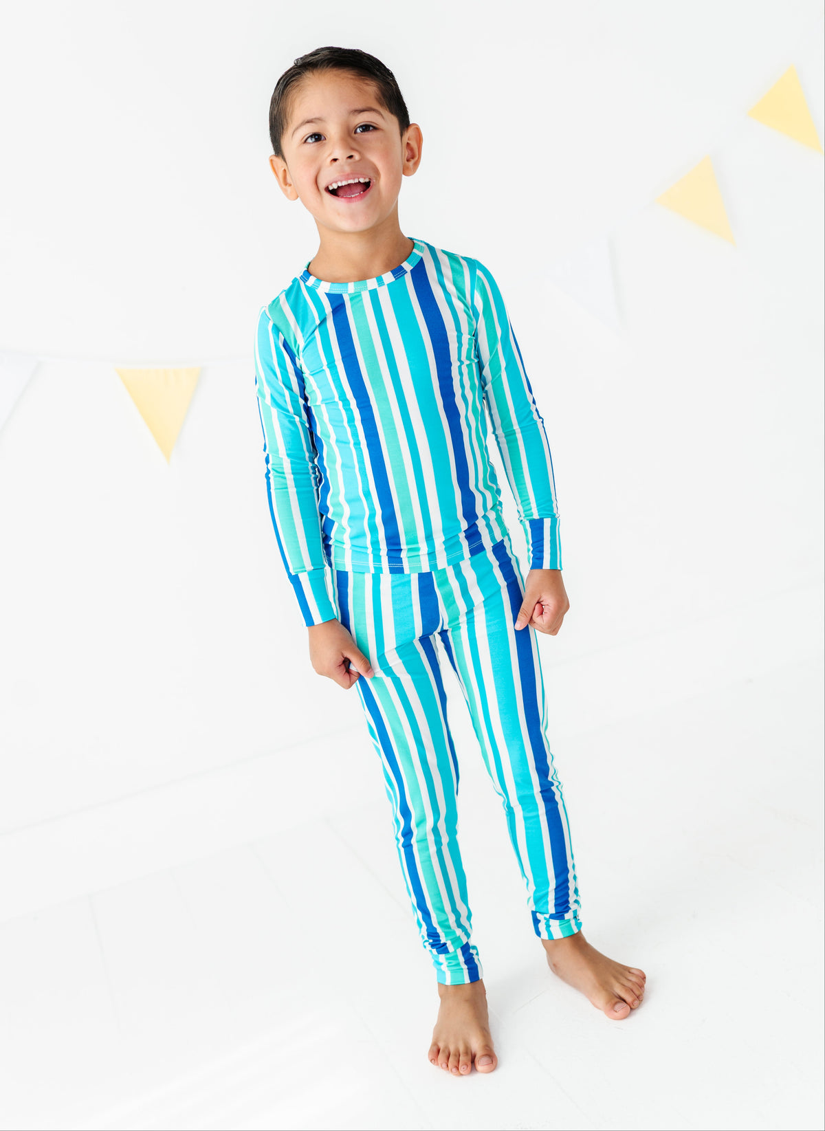 A young boy smiling and posing in a matching two-piece long-sleeve pajama set with vertical stripes in shades of blue, teal, and white, set against a clean white background with subtle yellow bunting.