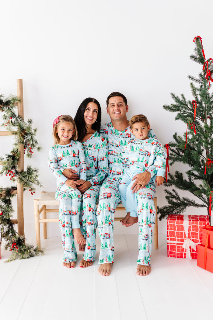 A family of four wearing matching Christmas-themed bamboo pajamas sits together on a white bench in a bright, holiday-decorated room. The pajamas feature a light blue background with a repeating pattern of green trees, red ornaments, and small red vehicles.