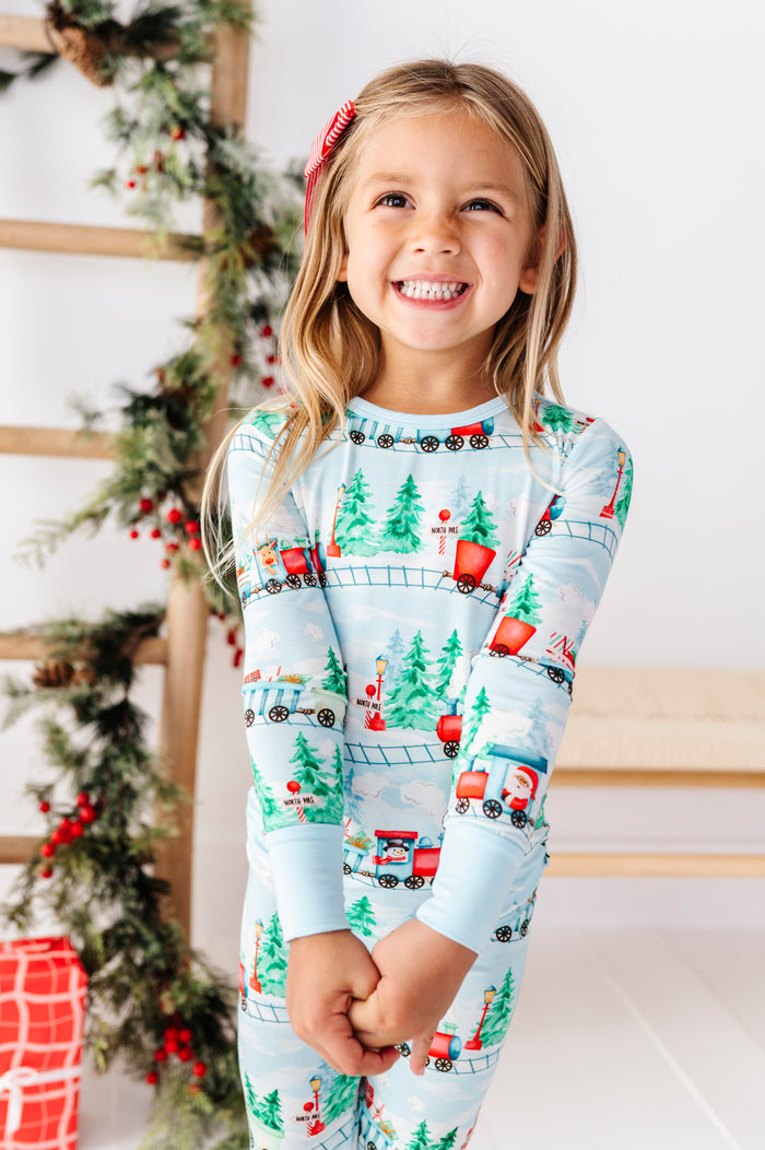 A young girl smiling while wearing a light blue long-sleeve bamboo pajama set with a Christmas train and Santa pattern, posed in front of a decorated holiday tree and wooden ladder.