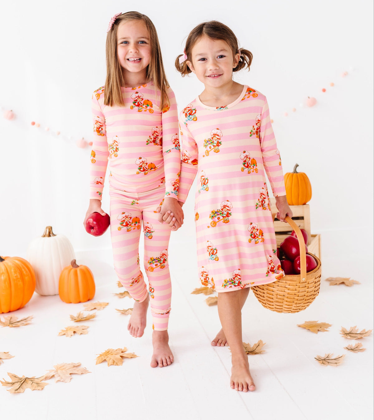 Two young girls wearing matching pink striped pajamas with a fall floral print walk barefoot in a studio decorated with pumpkins, leaves, and a basket of apples. A copyright watermark for Shortcake is visible at the bottom.