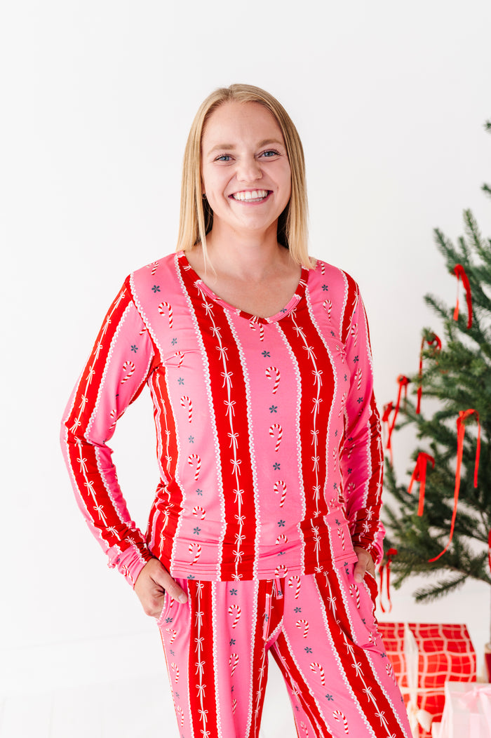 A smiling blonde woman wearing a festive pink and red long-sleeve bamboo pajama set with vertical candy cane and snowflake patterns, standing next to a decorated Christmas tree.