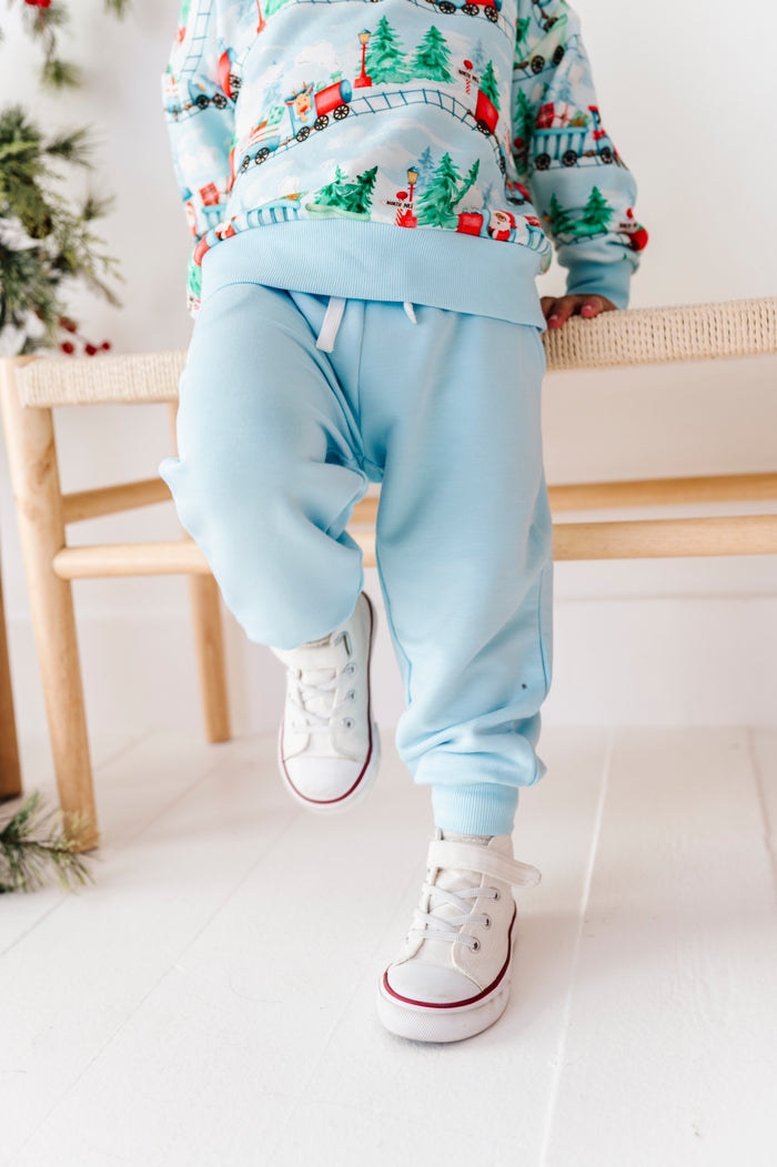 A child wearing a light blue sweatshirt and matching jogger pants with a Christmas train print, paired with white sneakers, sitting on a wooden bench in a bright room.