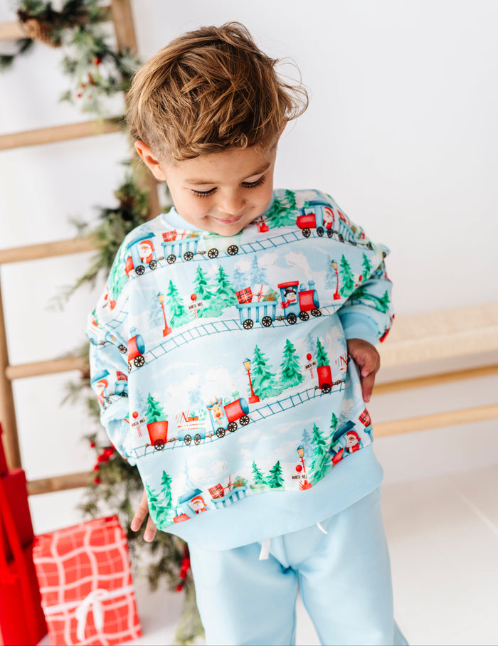 A young boy wearing a light blue crewneck sweatshirt with a festive Christmas train pattern and matching light blue sweatpants, standing in a holiday-themed room.