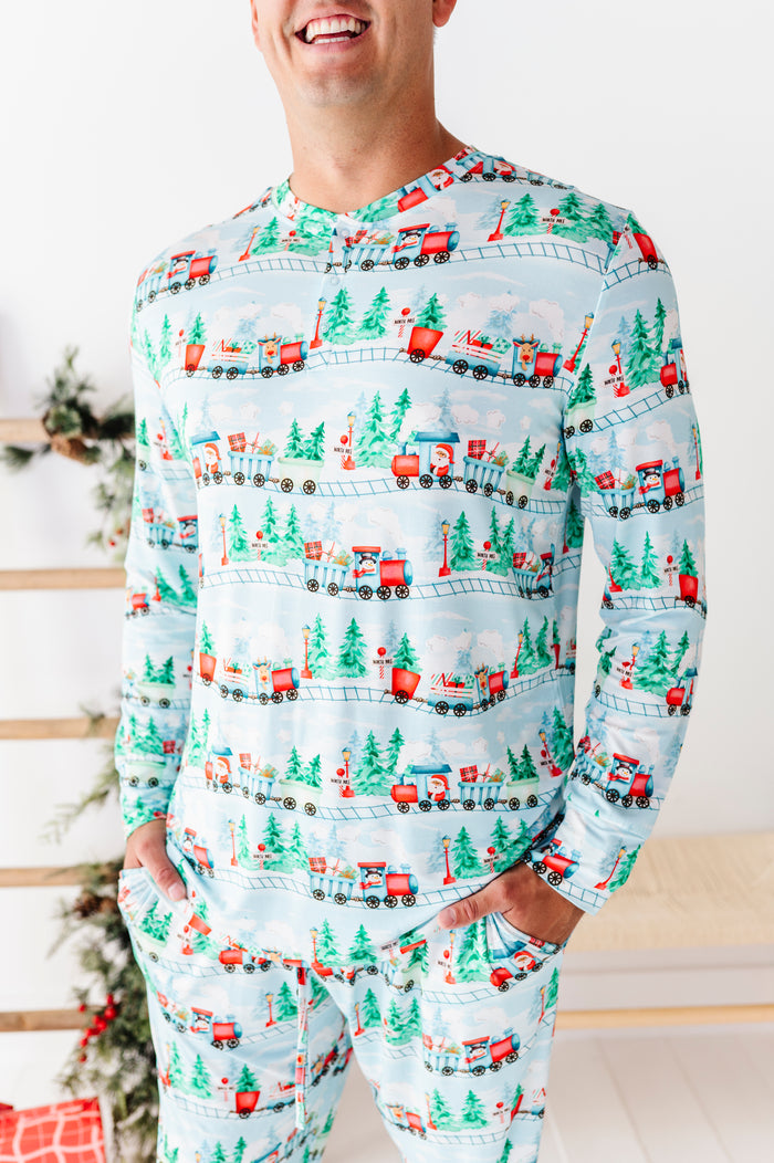 A man wearing a light blue long-sleeve bamboo pajama top and matching pants with a repeating Christmas train and pine tree pattern, standing indoors near a decorated holiday tree.