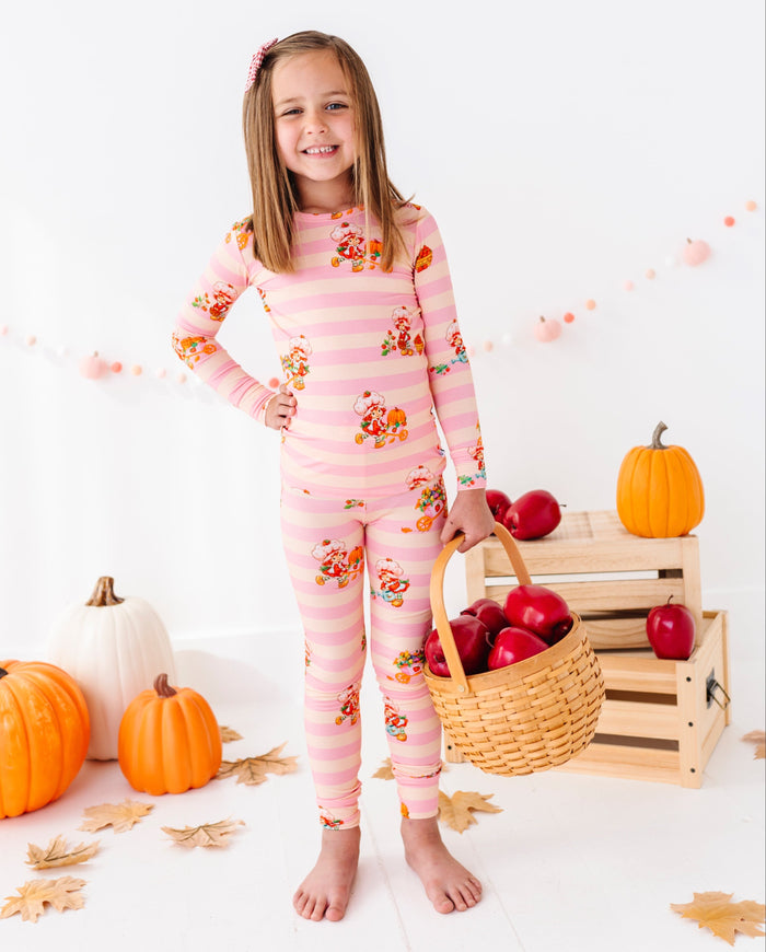 A young girl stands barefoot in a studio setting wearing a pink long-sleeve pajama set with horizontal stripes and a small floral print. She holds a wicker basket filled with red apples next to stacked wooden crates holding more apples and pumpkins.