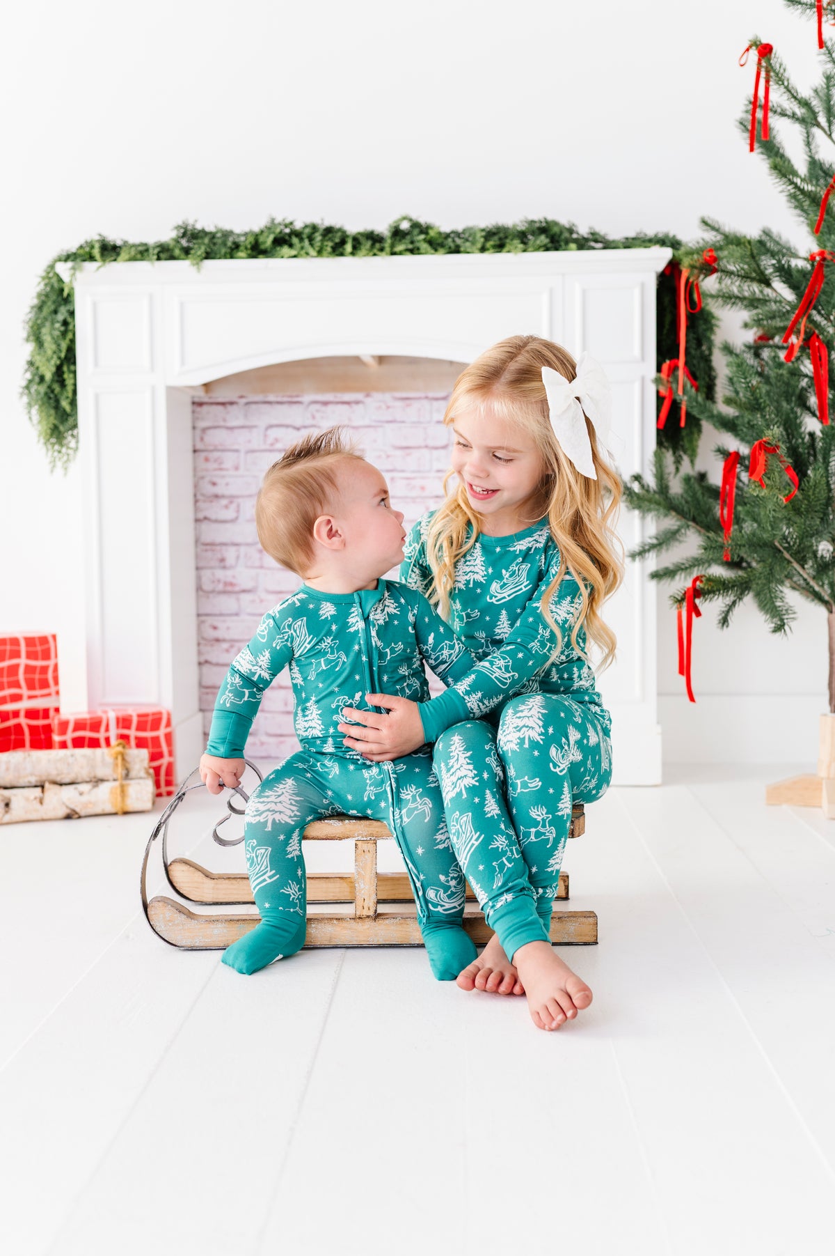 A little girl and her brother sitting on a wooden sleigh. They're wearing matching bamboo pajamas featuring vintage artwork including Santa and his reindeer