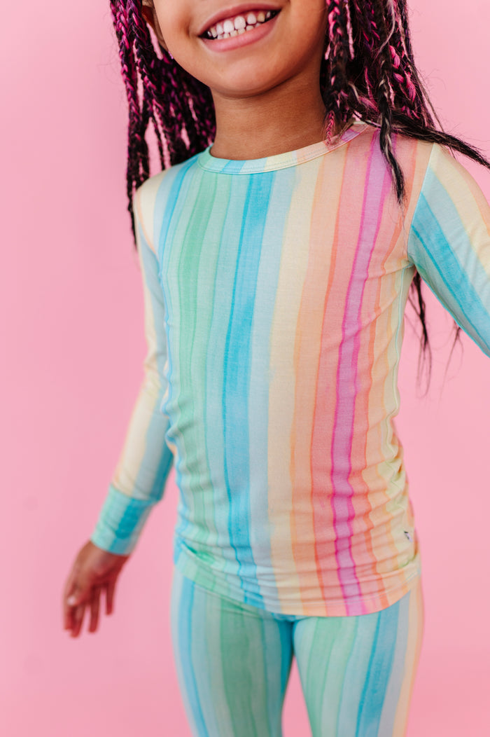 A girl wearing a rainbow striped bamboo pajama two-piece set against a pink background