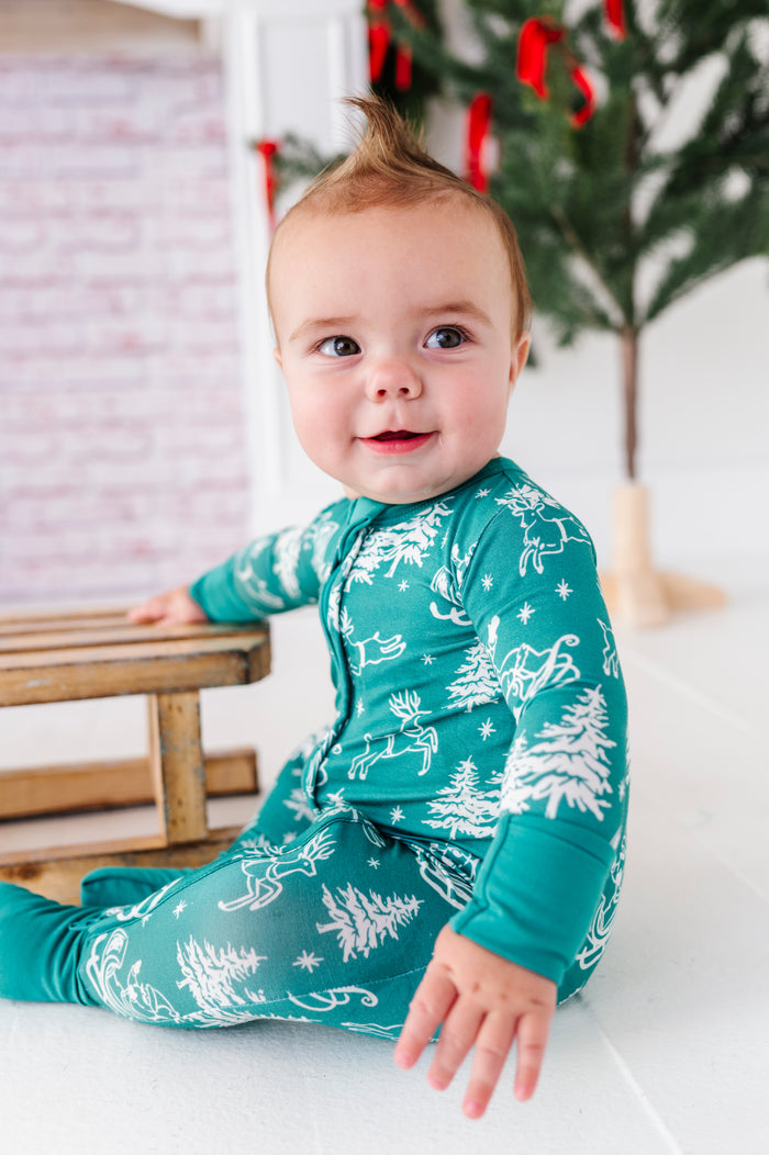 A little boy wearing a bamboo convertible romper and sitting in front of a fireplace. The romper features a vintage scenery print including Santa and his reindeer as they fly over the pine tree treetops