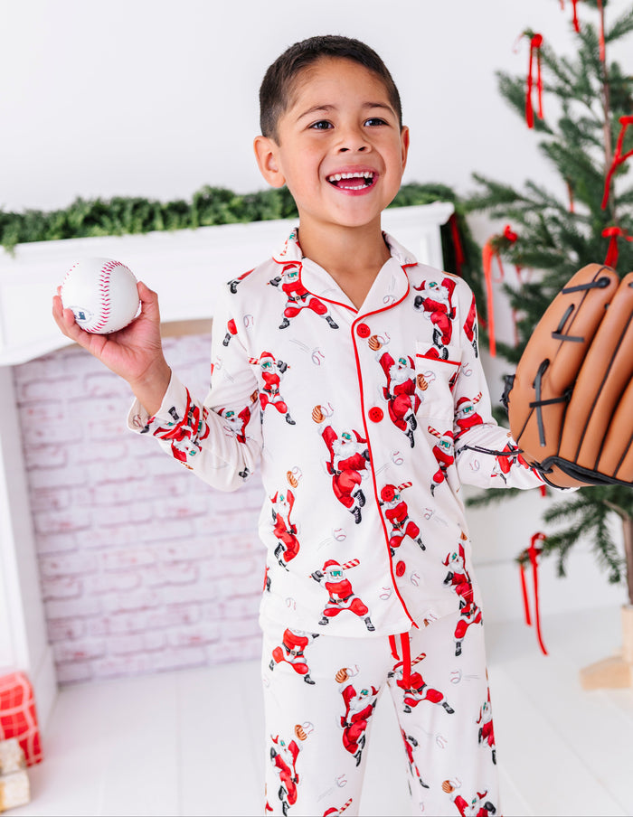 A little boy wearing a bamboo lounge set featuring Santa throwing baseballs and knocking them out of the park! He's holding a wooden bat while standing in front of a fireplace and a blurry Christmas tree