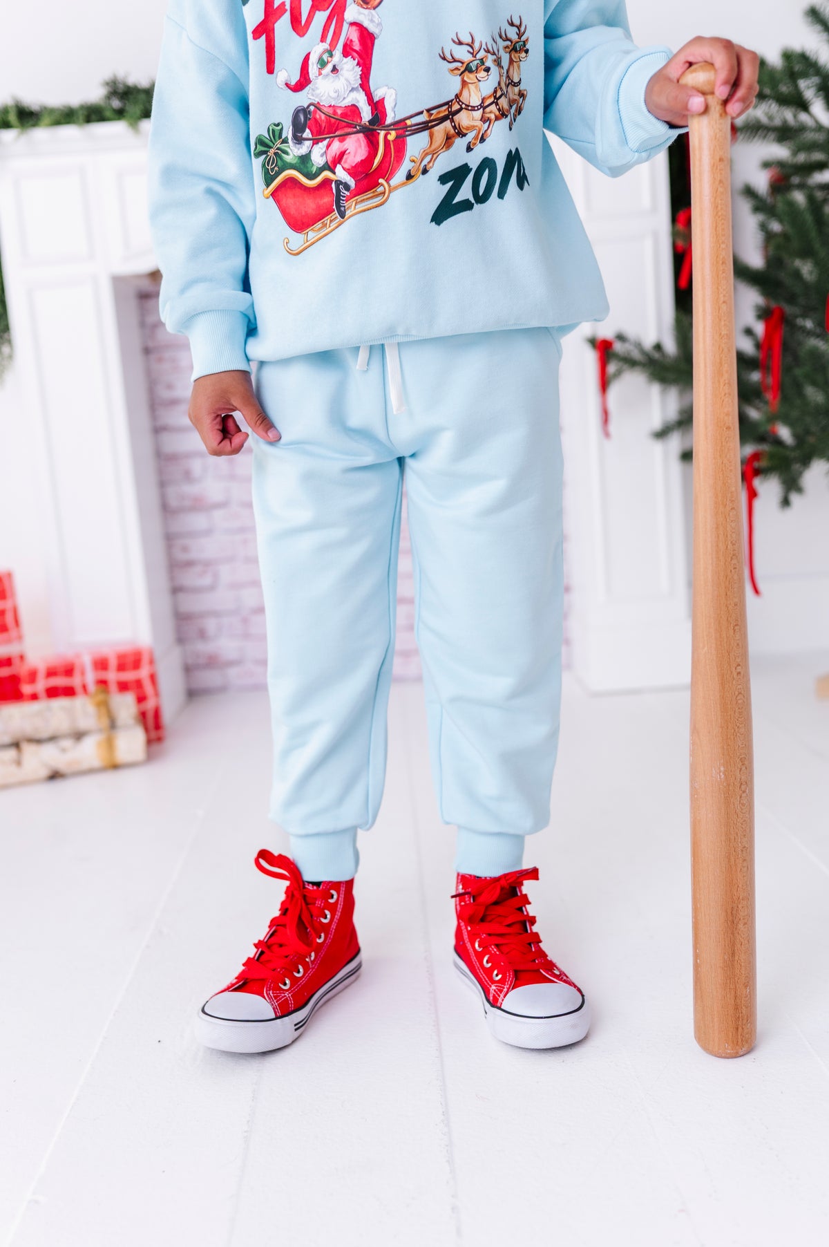 A young boy wearing a baseball crewneck with light blue bamboo joggers. He's holding a wooden baseball and standing in front of a fireplace