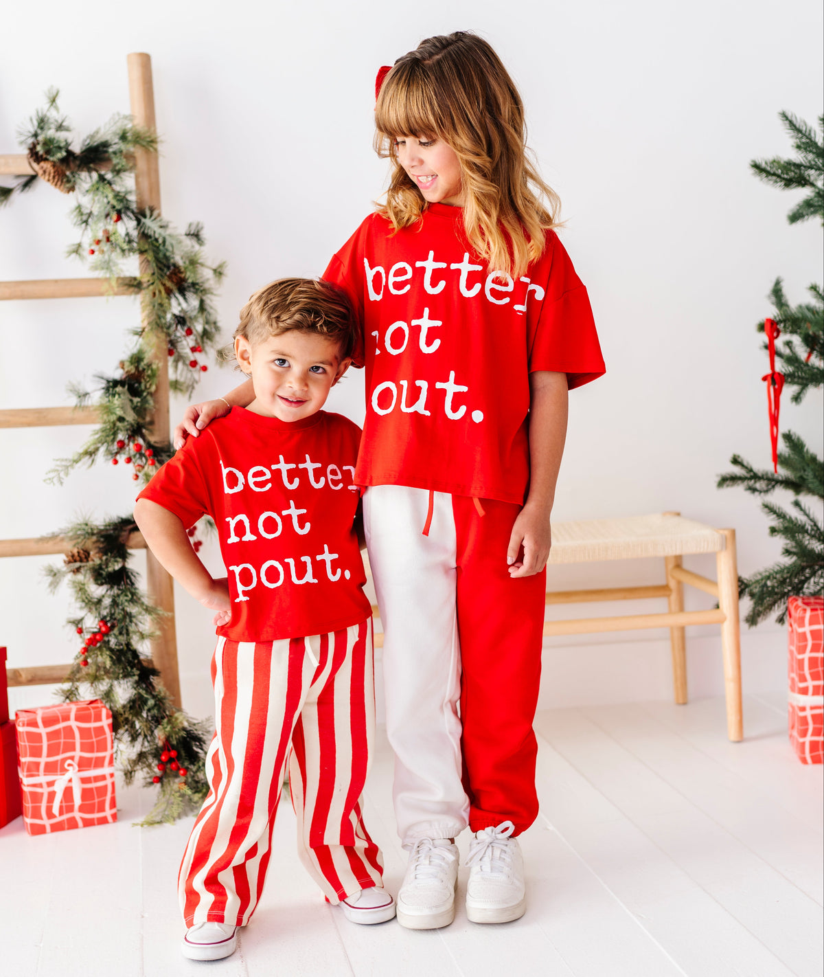 Two children standing together in a bright white room decorated for Christmas, wearing matching red graphic sweatshirts that say 'better not out.' and red-and-white striped pants.