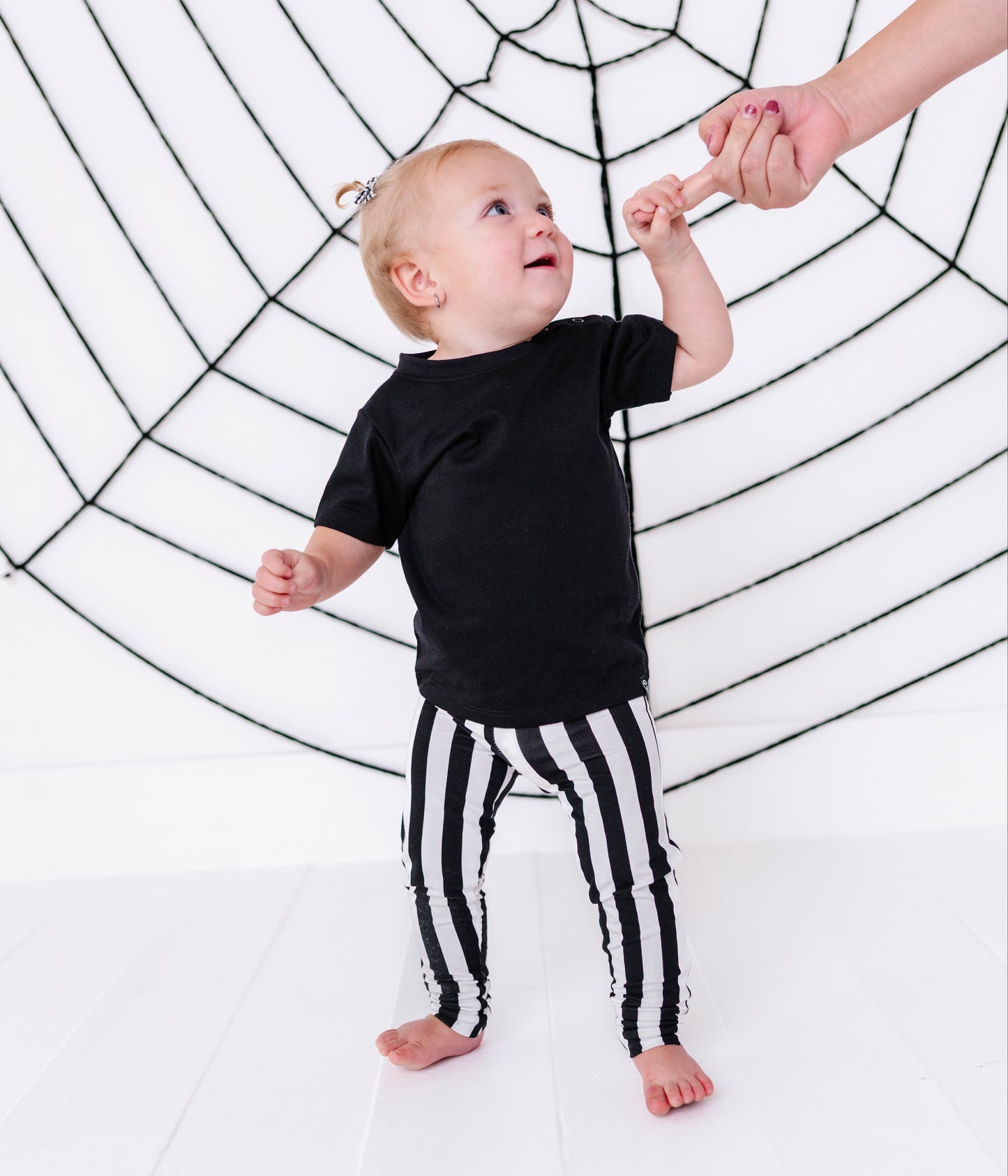 A toddler standing on a white floor in front of a large black spiderweb backdrop, wearing a black short-sleeve top and black-and-white vertically striped pants, reaching up to hold an adult hand.