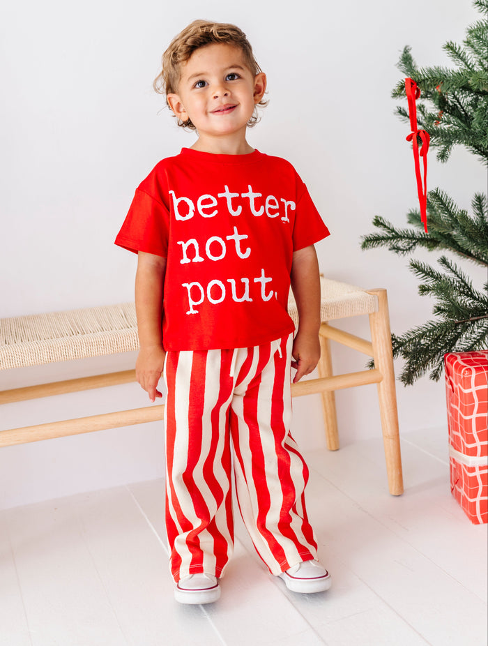 A toddler stands in a bright indoor setting wearing a red short-sleeve T-shirt with white text reading 'better not pout' and red-and-white vertically striped pants. A wooden bench, white wall, and part of a Christmas tree with a wrapped gift are visible in the background.