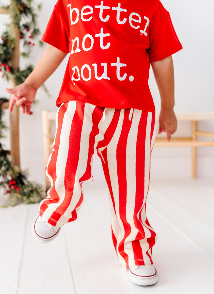 A child wearing a bright red short-sleeve t-shirt with white text reading 'better not bout.' and red-and-white vertically striped pants, shown from the back in a bright indoor setting.