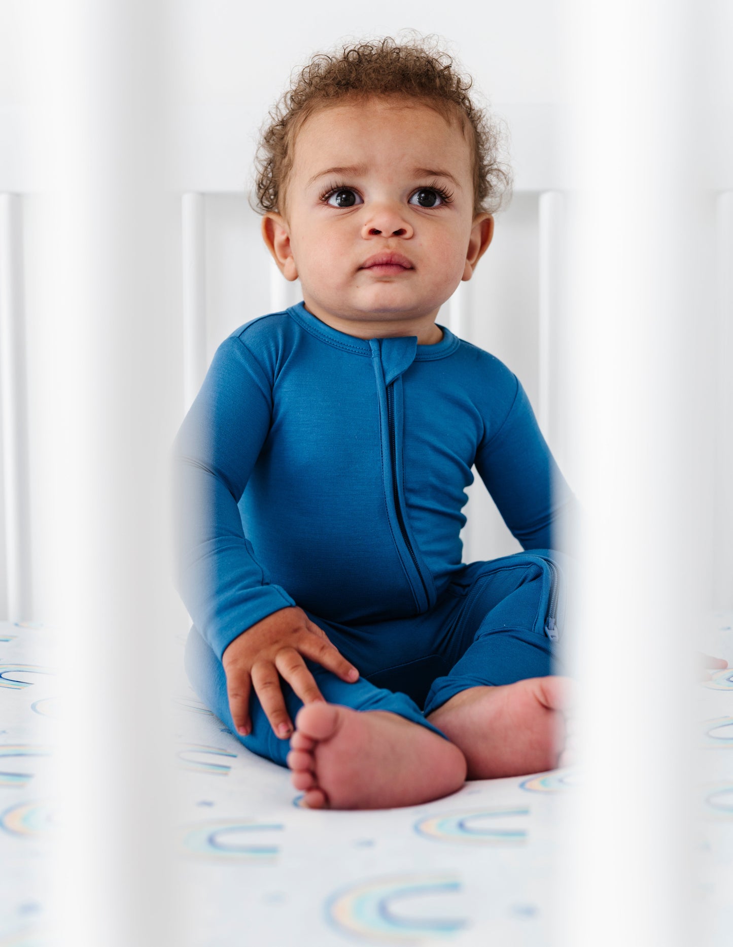 Baby in a blue bamboo zip romper sitting in a crib with a white background