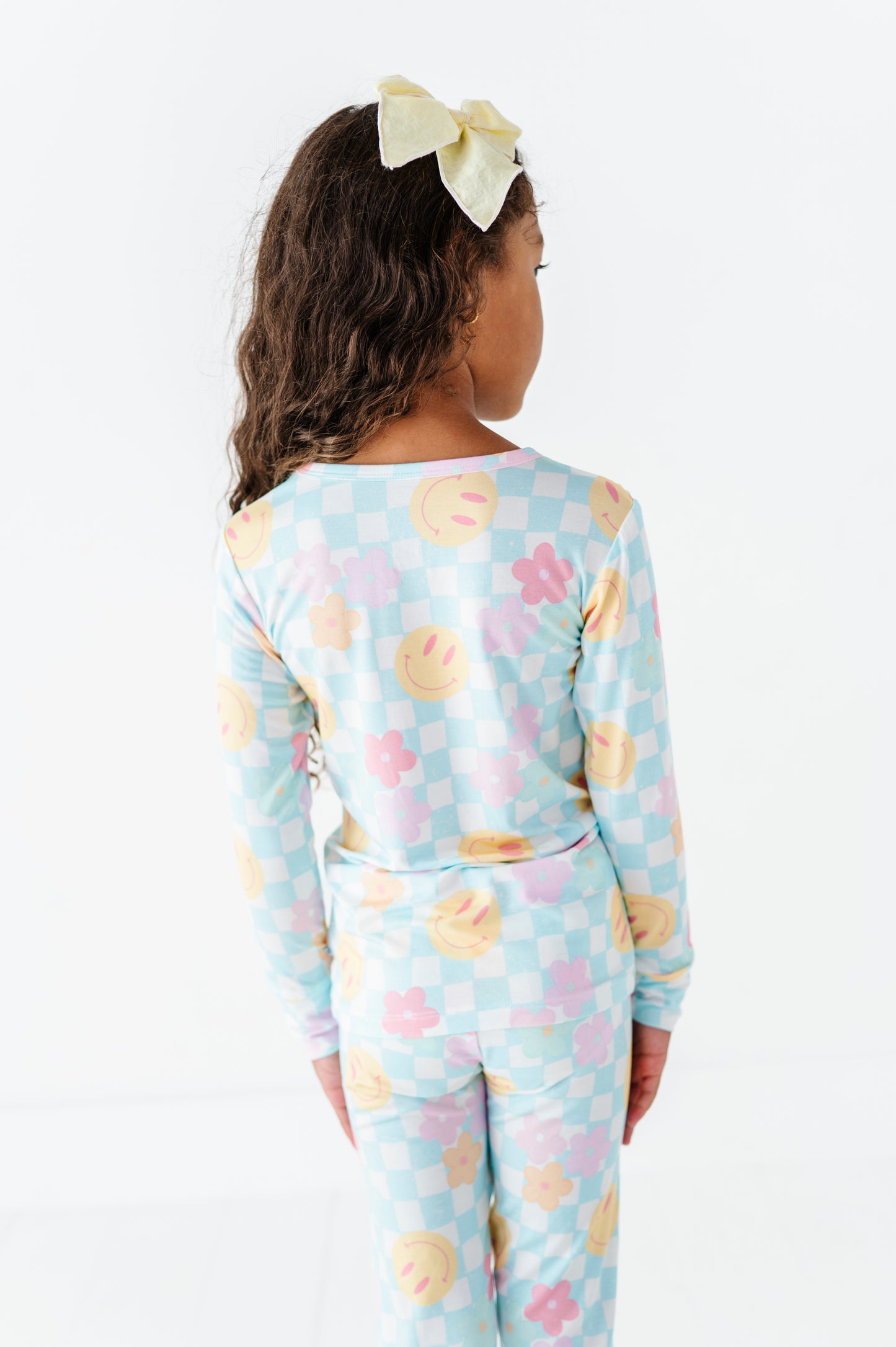 A child model shown from the back wearing a light blue long-sleeve pajama set with a pastel checkerboard pattern and pink, yellow, and orange smiley face motifs. The model has dark brown curly hair and wears a pale yellow bow headband against a clean white background.