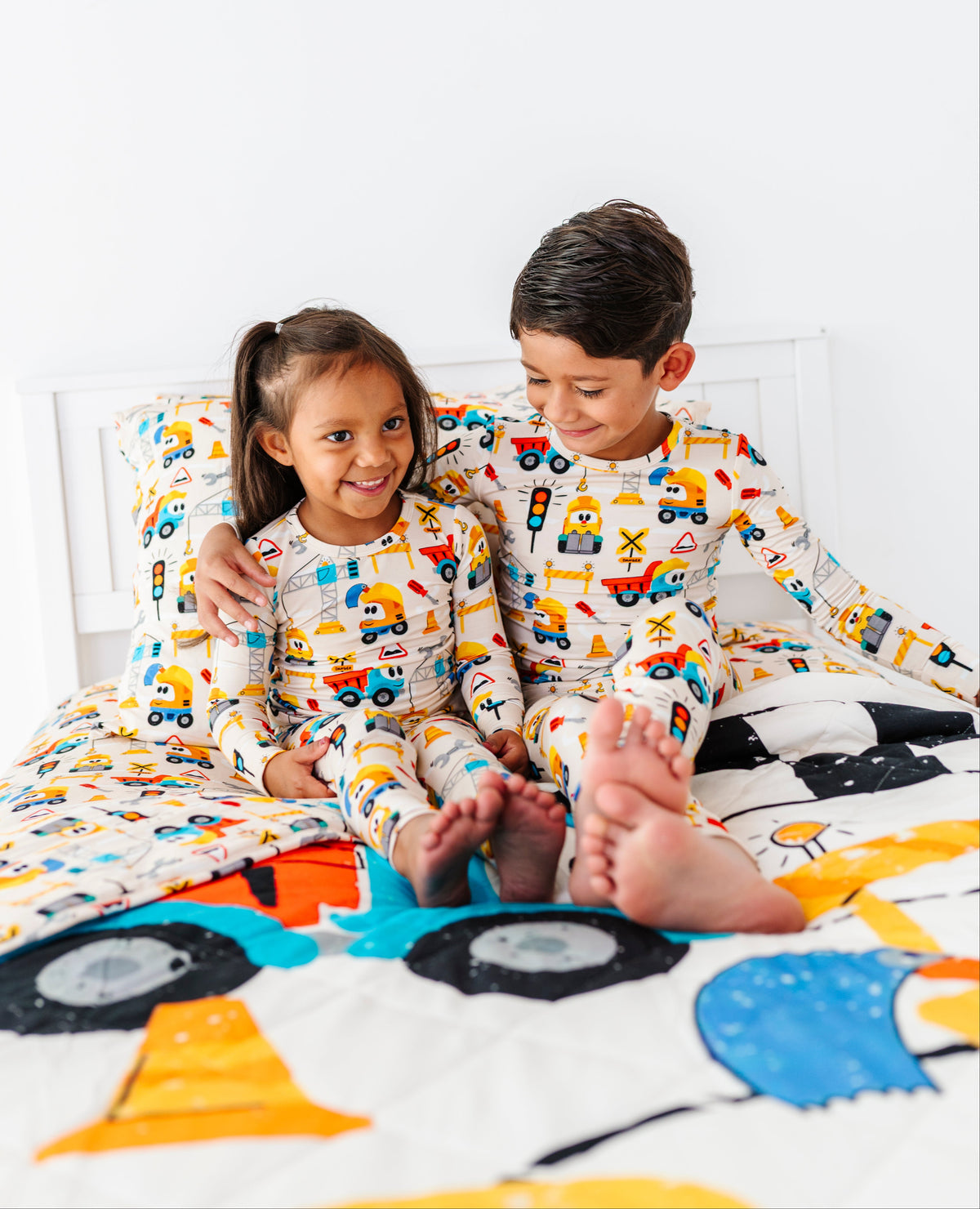 Two children sitting on a bed wearing matching white long-sleeve pajamas with a colorful construction vehicle print, with a matching quilt in the foreground.