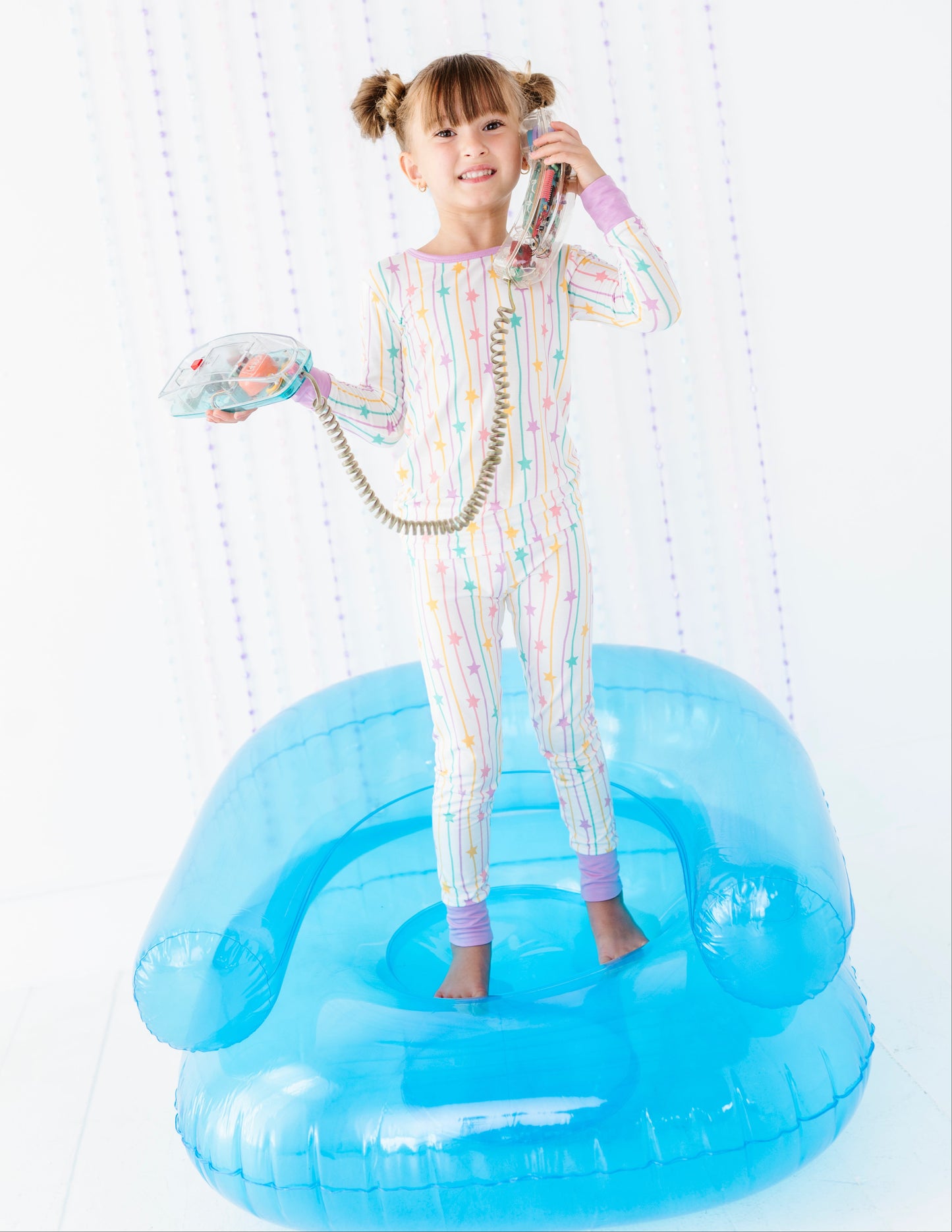 A young girl with pigtails stands on a translucent blue inflatable pool float, wearing a pastel rainbow-striped one-piece pajama set and holding a toy telephone receiver and handset against a white shower curtain background.