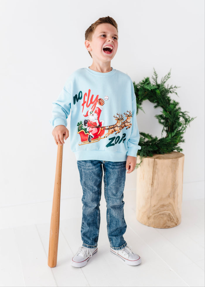 A young boy stands in a bright white room holding a baseball bat and wearing a blue crewneck that says "no fly zone"