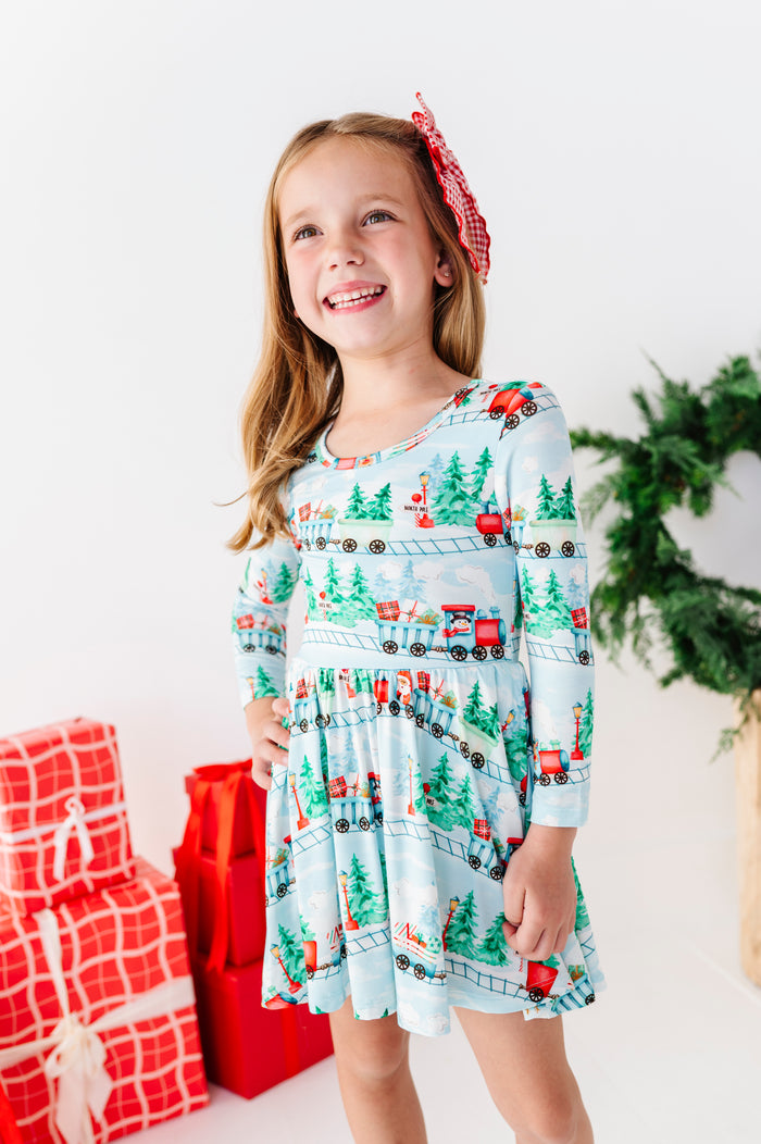 A young girl smiling and wearing a light blue long-sleeve bamboo dress with a Christmas train print, standing in a bright studio with wrapped red gifts, a green wreath, and a wooden stool.