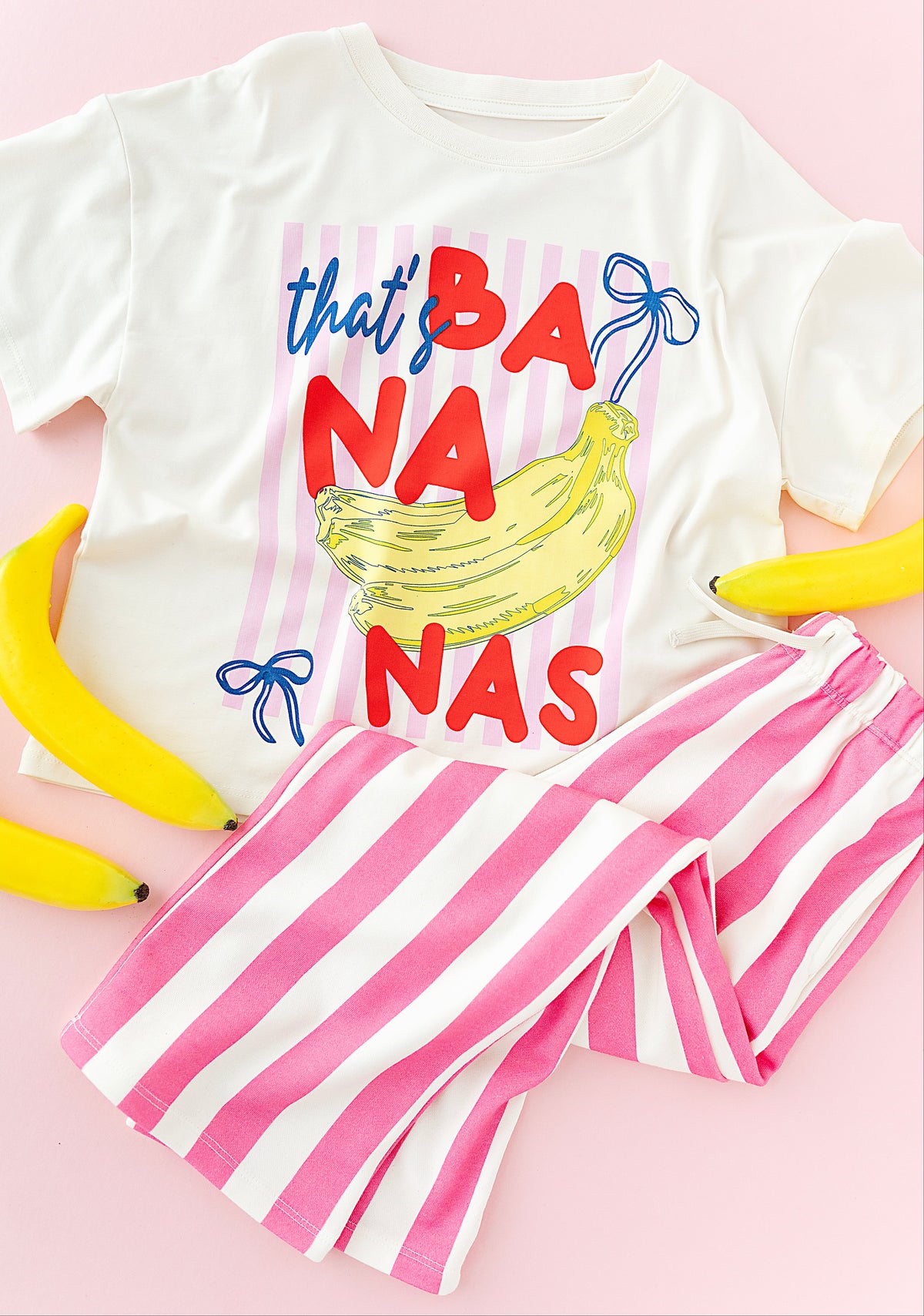 A white short-sleeve T-shirt with a colorful graphic reading 'that's BANANA'S' in blue and red lettering around a yellow banana illustration, paired with pink and white striped shorts on a light pink background with decorative bananas around them.