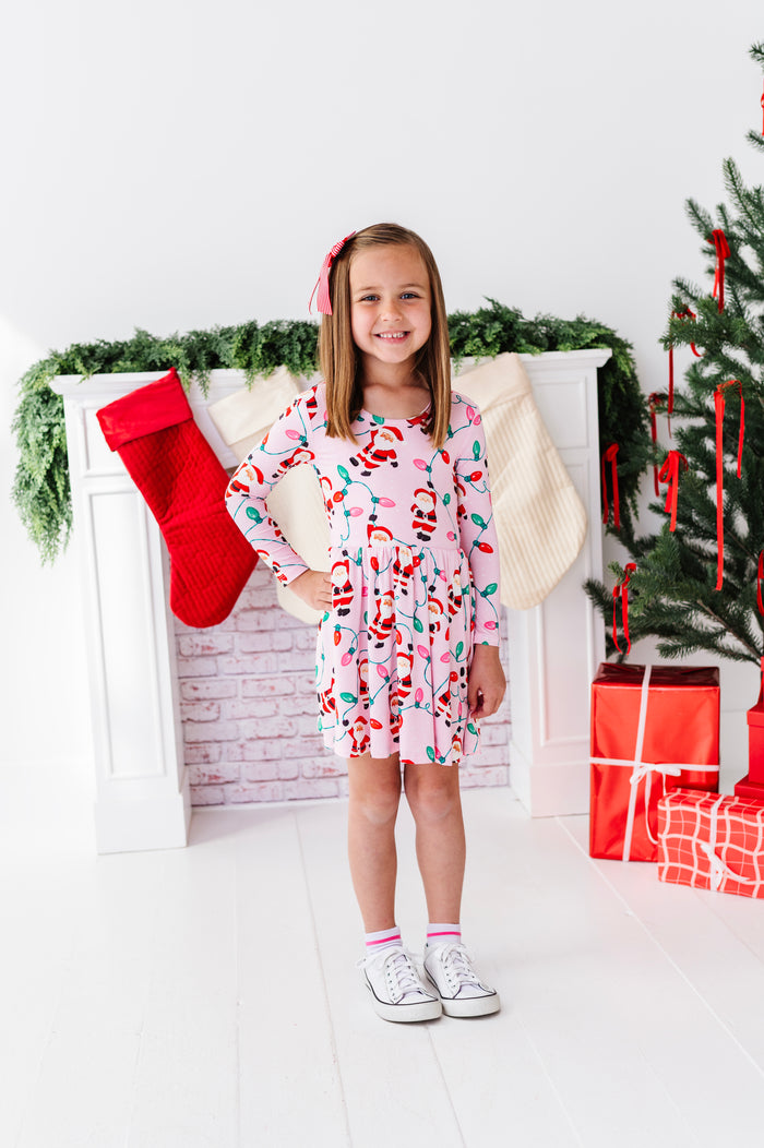A young girl stands in a holiday-themed room wearing a pink long-sleeve bamboo dress with a colorful Santa Claus pattern. The background features a white fireplace mantel with a red stocking, green garland, and a Christmas tree with wrapped presents.