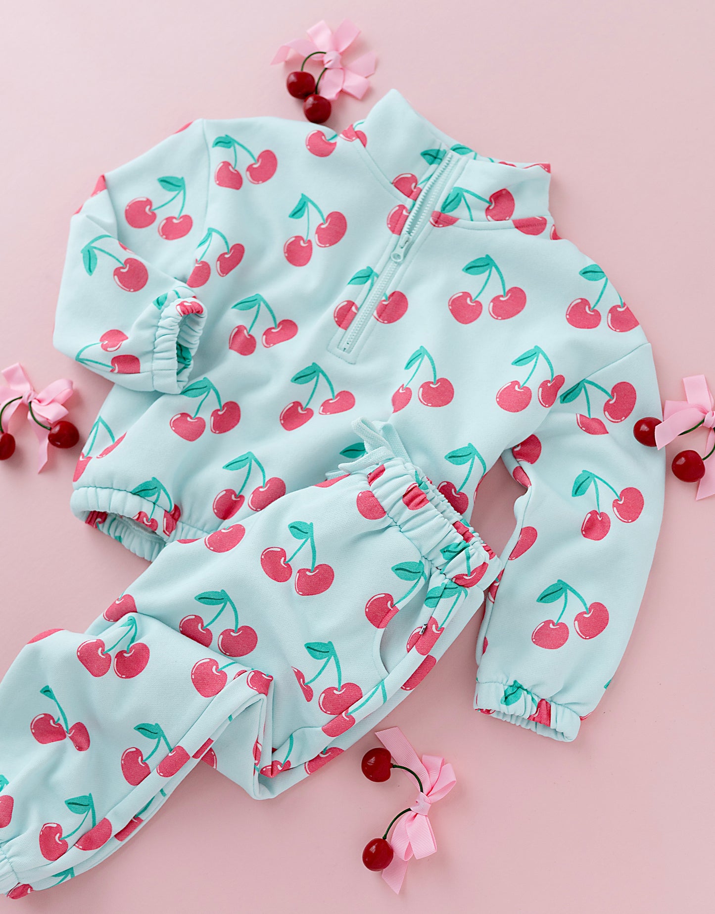 A baby two-piece outfit set consisting of a light blue zip-up jacket and matching pants, both covered in a red cherry print, arranged on a pink background with decorative cherry props.