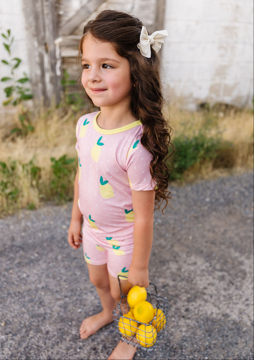 Young girl in pink lemon-themed pajamas holding lemons outdoors.