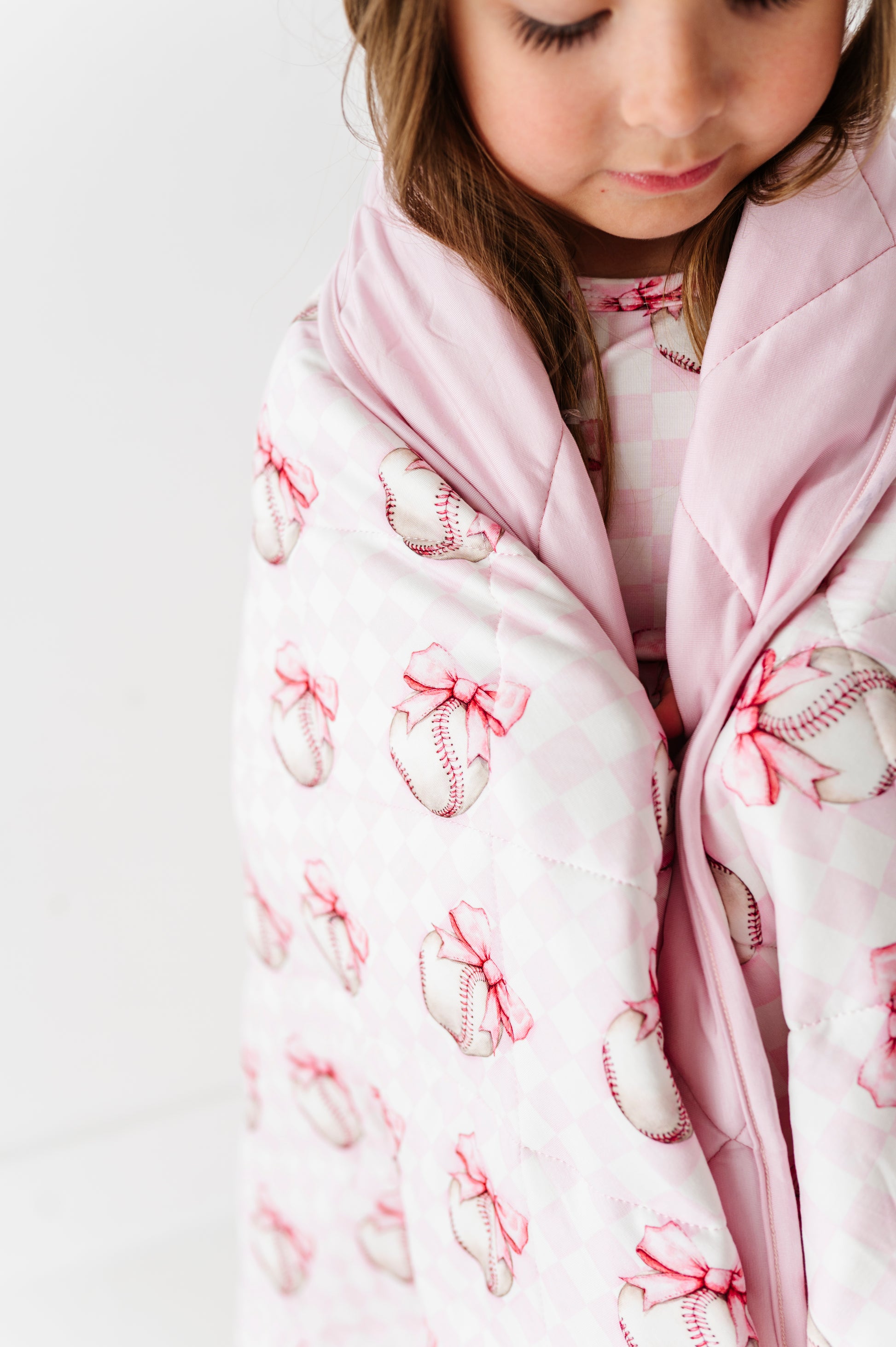 Child wrapped in a pink bamboo blanket with baseball and bow pattern on a white background