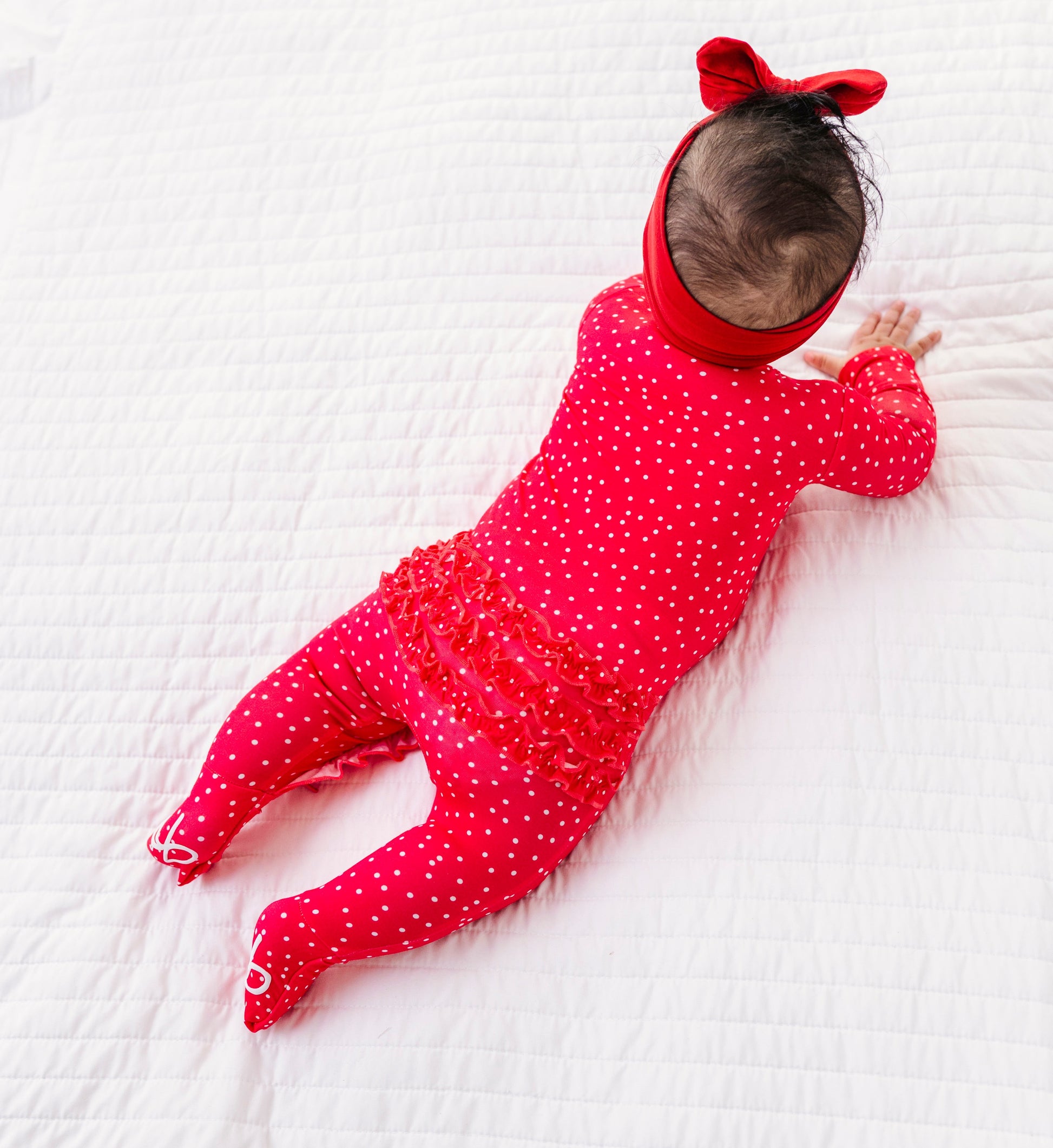 Baby in a red polka dot outfit with a matching headband lying on a white textured surface.