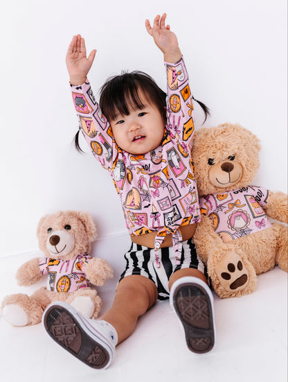 A toddler girl sitting on a white floor with her arms raised, wearing a pink long-sleeve crop top and black-and-white striped shorts, surrounded by two teddy bears wearing matching outfits.