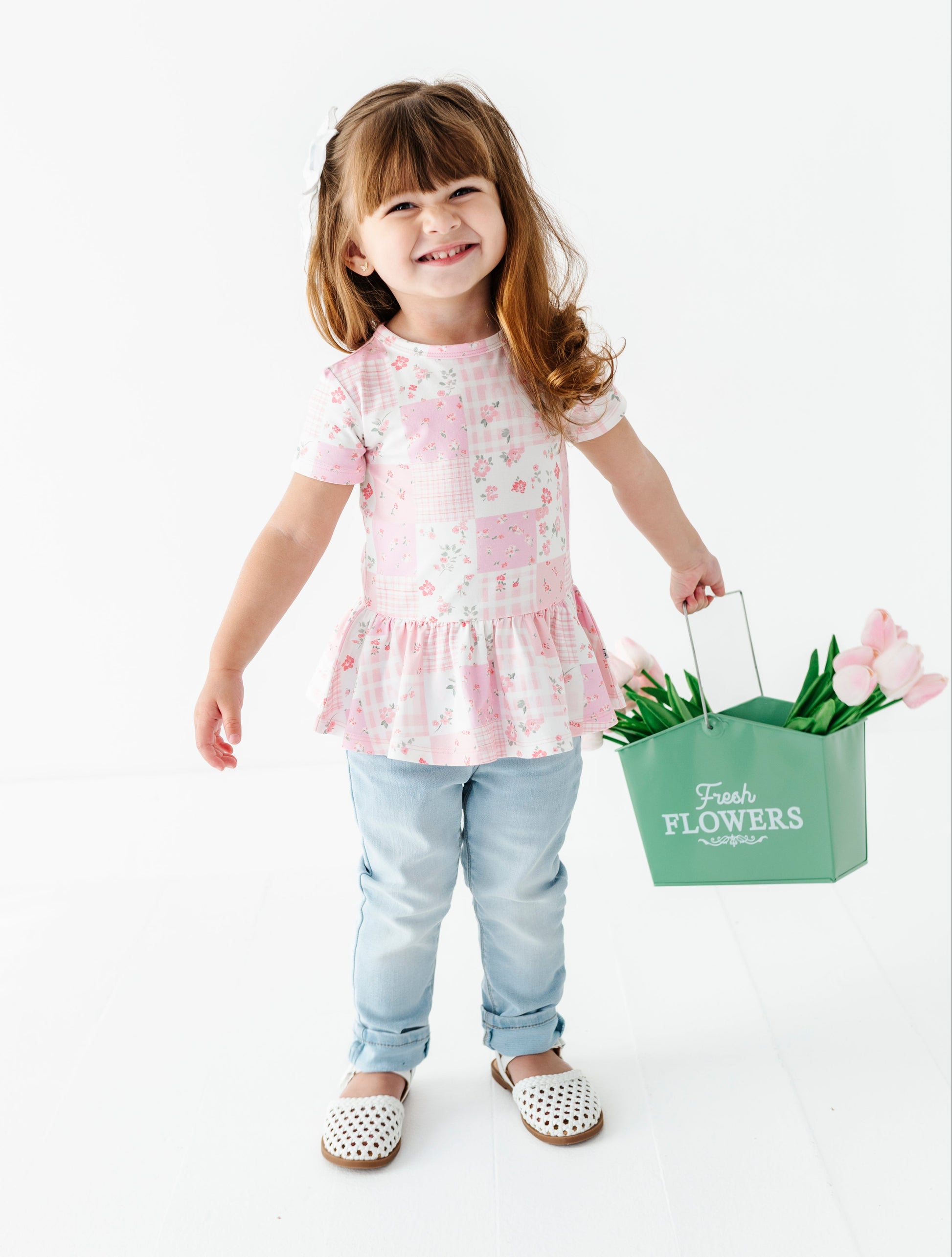 Child holding a green 'Flowers' basket with tulips wearing a bamboo-cotton peplum blouse on a white background