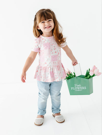 Child holding a green 'Flowers' basket with tulips wearing a bamboo-cotton peplum blouse on a white background