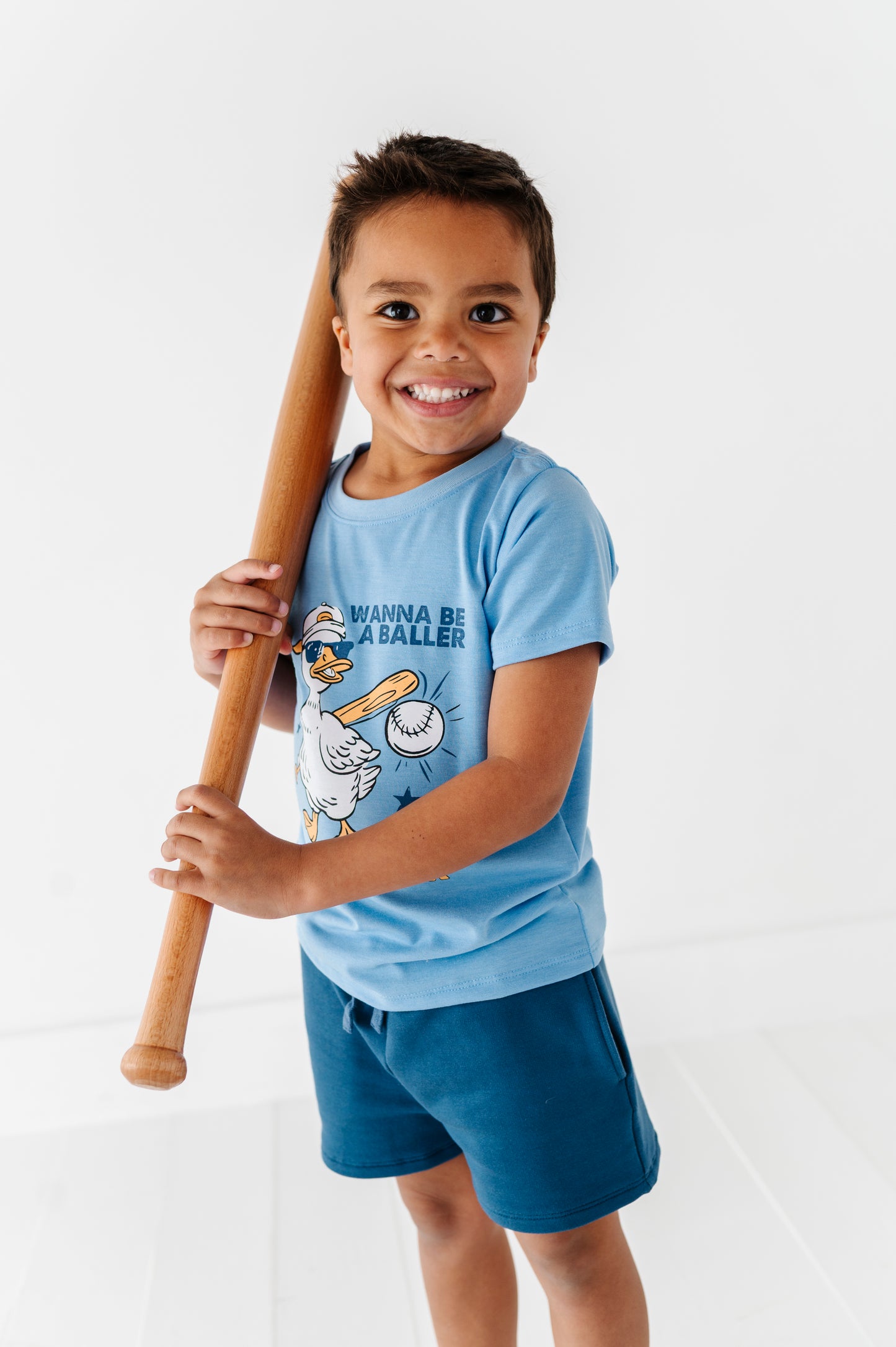 Child wearing a blue baseball t-shirt with a graphic design and holding a baseball bat on a white background