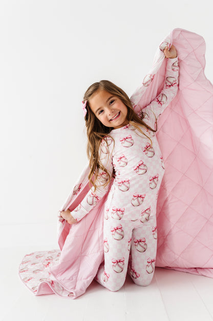Young girl in pink bamboo pajamas with baseball and bow pattern holding a pink quilt on a white background