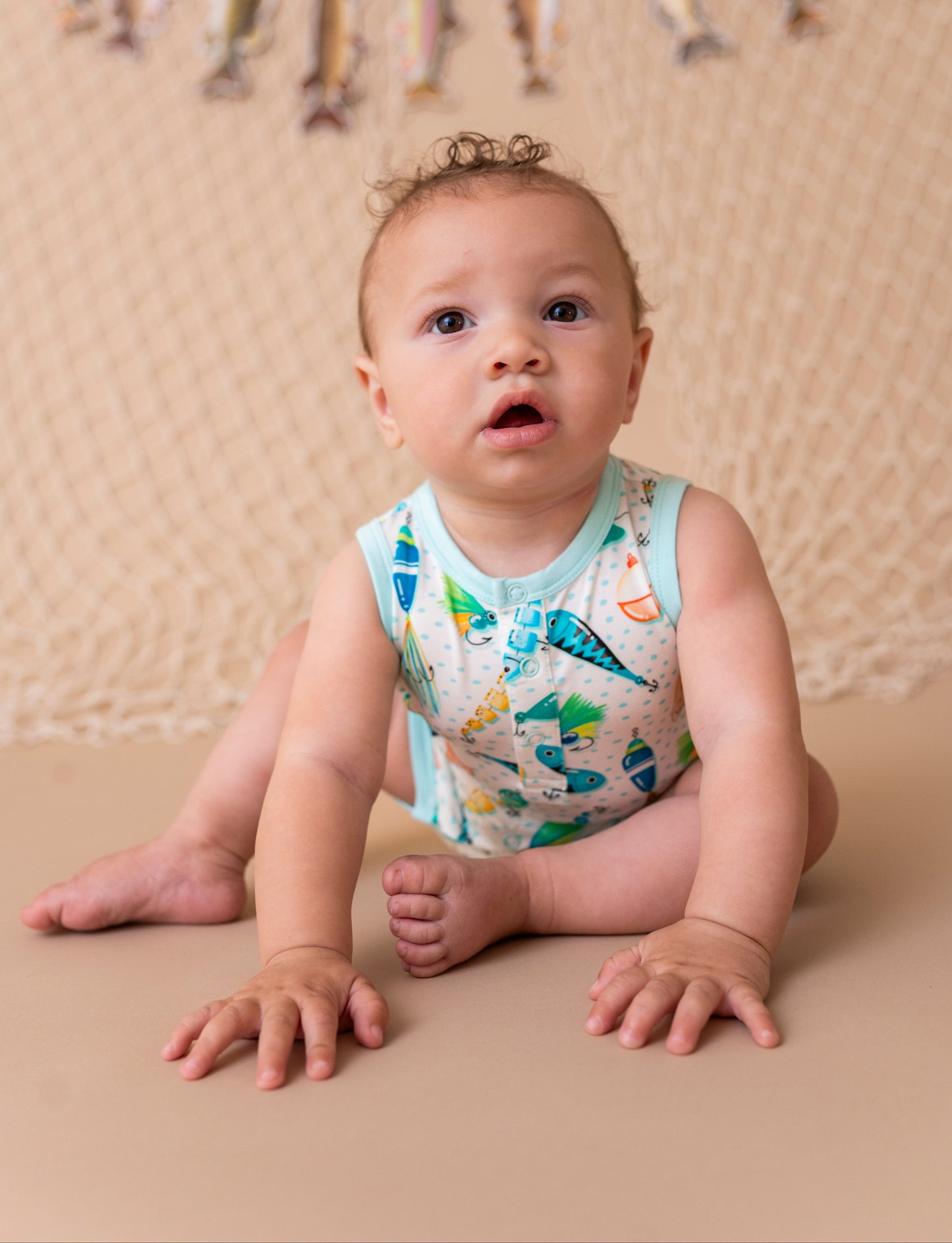 Baby sitting on a beige surface wearing a colorful sleeveless shirt with fishing lure print, against a beige backdrop with decorative elements.