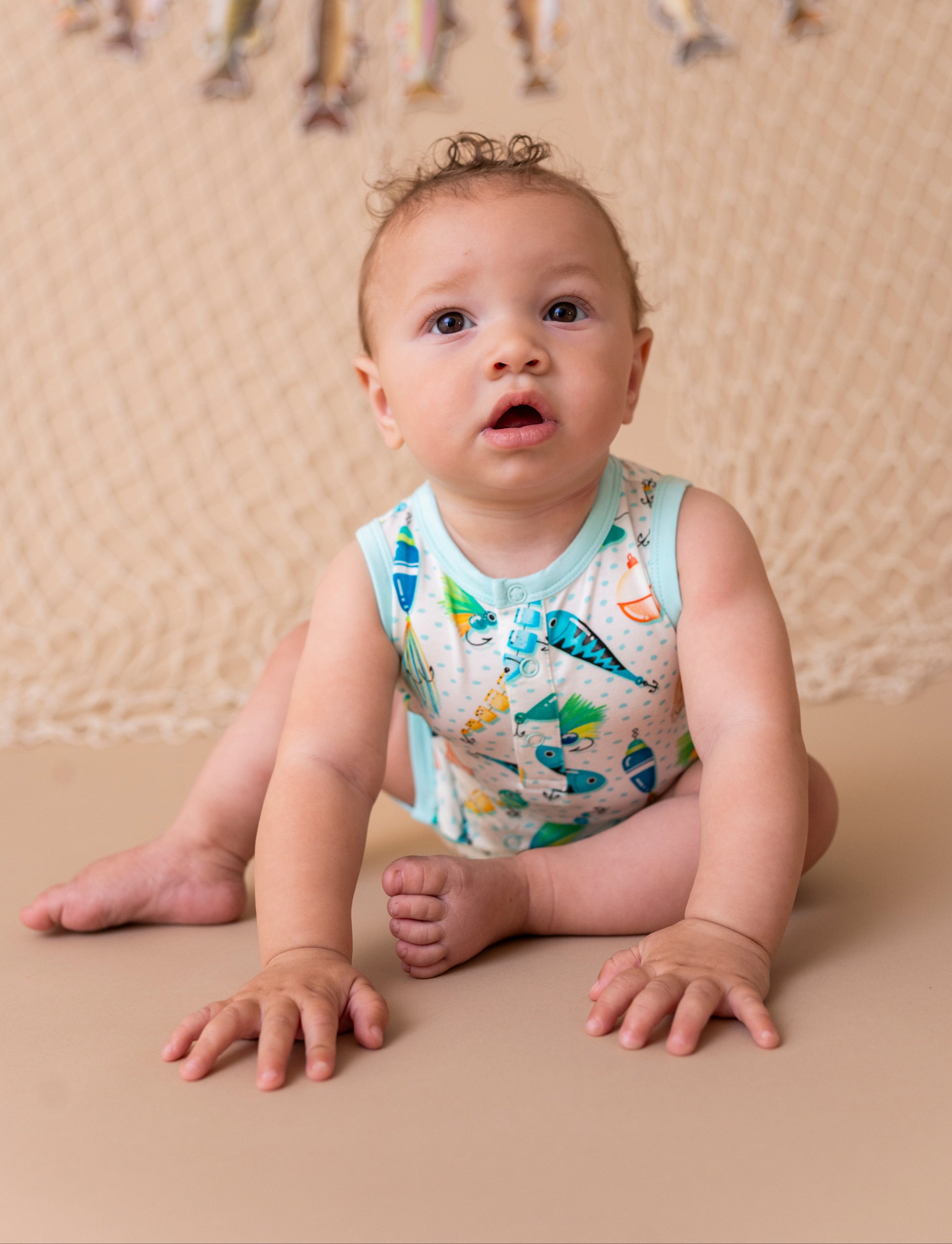 Baby sitting on a beige surface wearing a colorful sleeveless shirt with fishing lure print, against a beige backdrop with decorative elements.