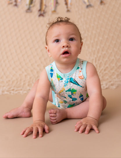 Baby sitting on a beige surface wearing a colorful sleeveless shirt with fishing lure print, against a beige backdrop with decorative elements.