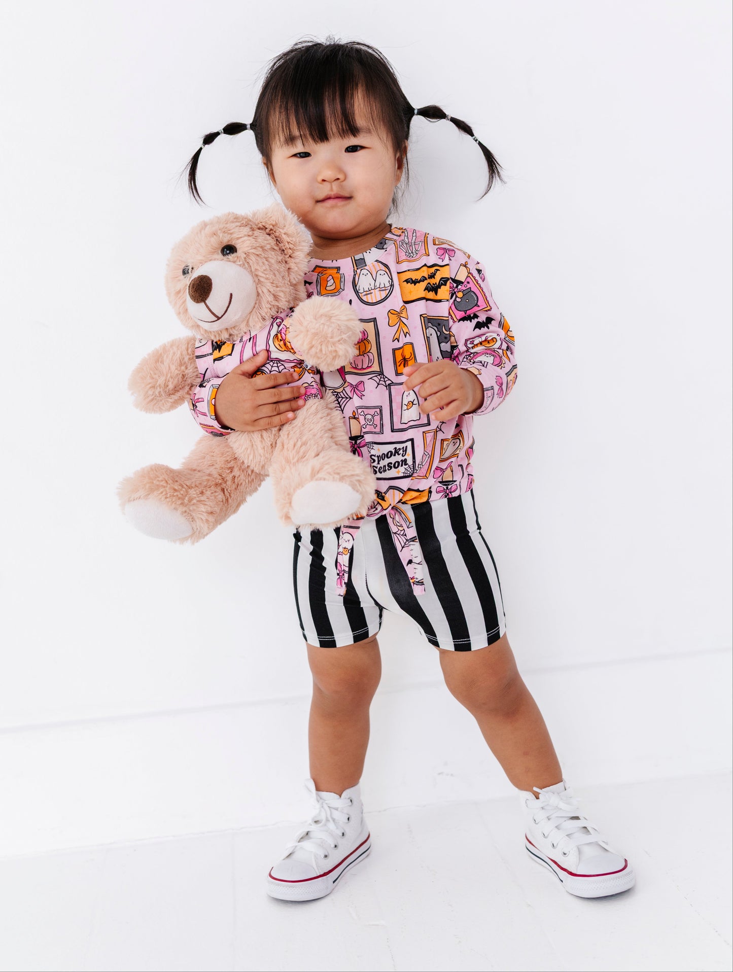 A toddler girl with black pigtails stands against a white studio background, holding a light brown teddy bear. She is wearing a pink long-sleeve top with a multicolor graphic print, black and white vertically striped shorts, and white low-top sneakers.